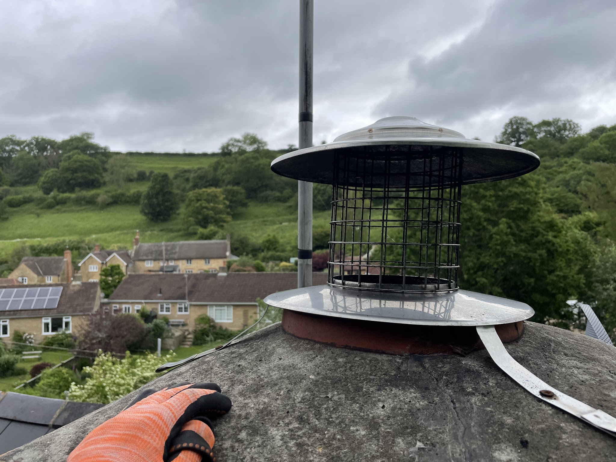 A stainless chimney cowl with a bird guard on a chimney pot, with a Somerset village and hills behind